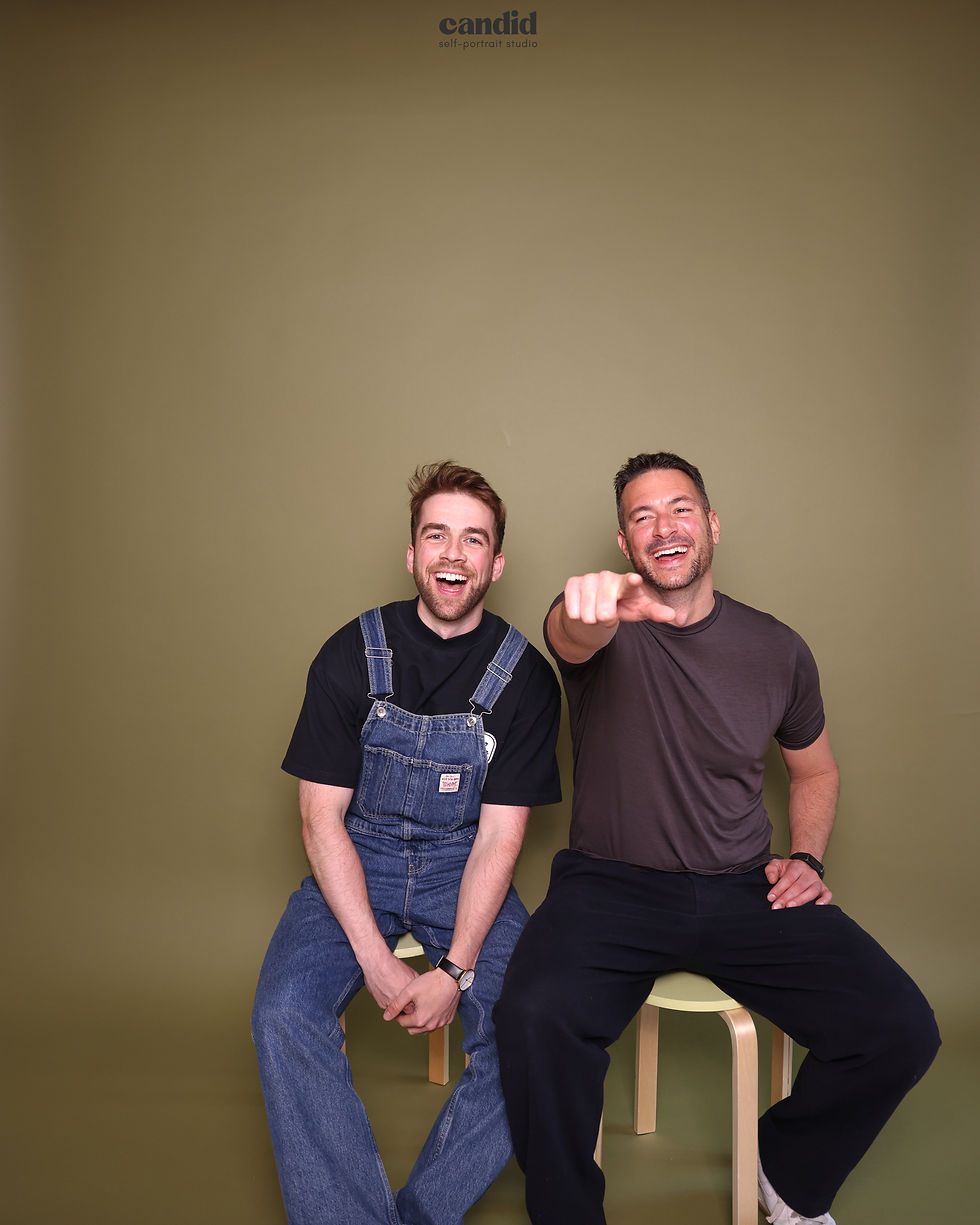 Two smiling men sit on stools in a studio; one points at the camera. Candid self-portrait studio text above.