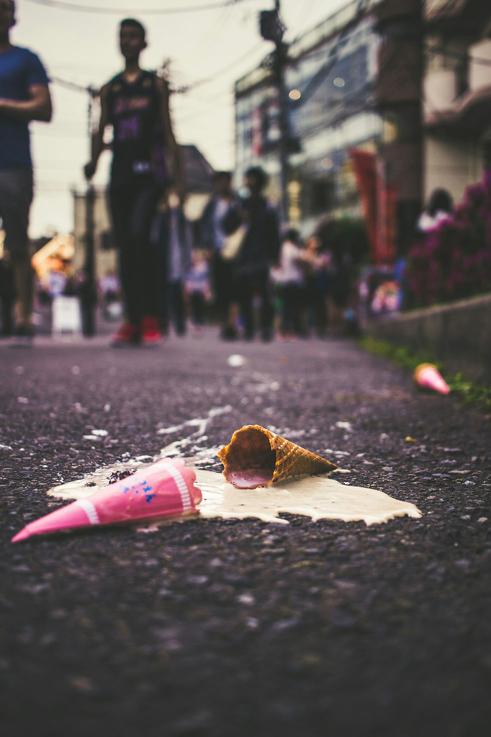 Pink ice cream cone spilled on wet asphalt as blurred pedestrians pass in a city street.