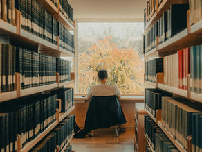Person seated in a quiet library aisle between tall bookcases, facing a window with autumn trees outside.