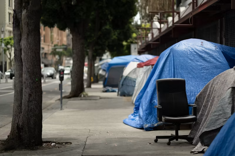 Row of blue and gray tents on a city sidewalk, with a black office chair beside them under trees and apartment buildings.