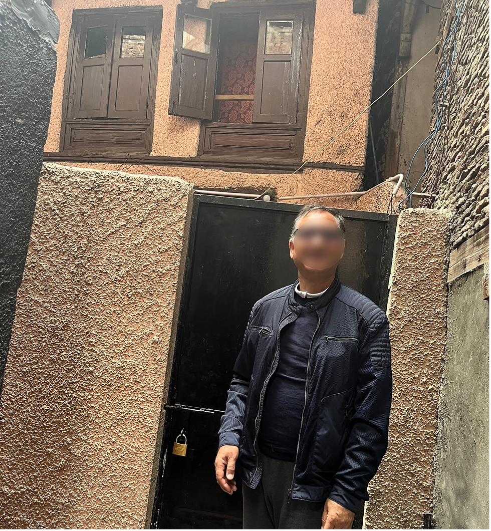 A man with a blurred face is standing outside a black metal door to a light brown-coloured house.