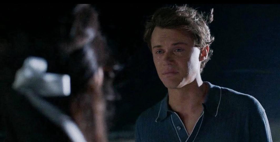 Tearful young man (Conrad Fisher) in a dark blue shirt faces a blurred figure in a dim room, looking sad and tense.