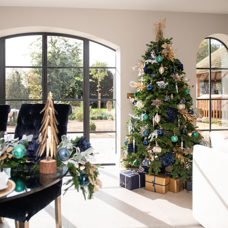 Festive room with decorated Christmas tree in gold, blue, and green. Presents below. Sunlit, large windows reveal greenery outside. Cozy ambiance.