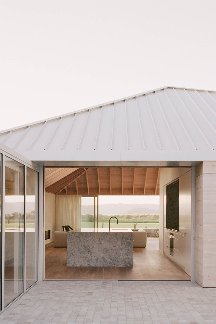 Modern home interior with open sliding glass doors, a marble kitchen island, light wood floors, and a scenic view of hills in soft daylight.