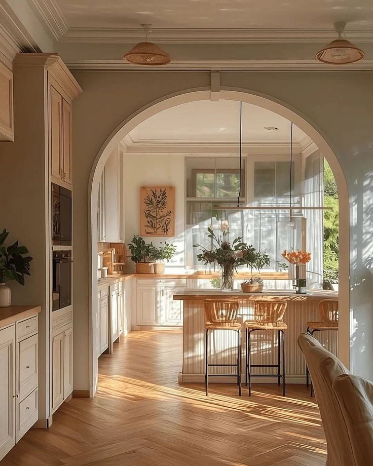 Airy kitchen with warm tones and sunlight streaming through large windows. Wooden floors, wicker chairs, and flowers create a serene mood.