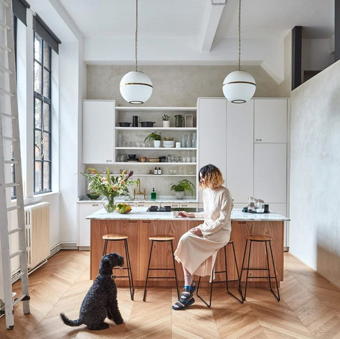 Woman in a modern kitchen sits on a stool at a wooden island, reading. A black dog sits nearby. Bright space with plants and pendant lights.