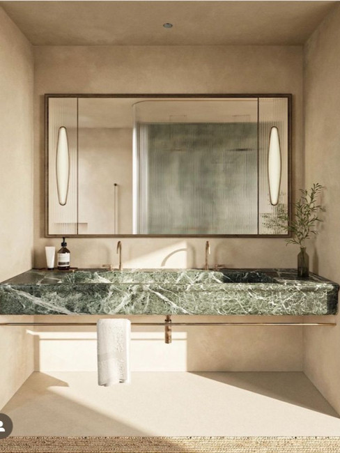 Elegant bathroom with a large mirror, green marble sink, gold faucets, and towel. Beige walls, minimal decor with a plant on the right.
