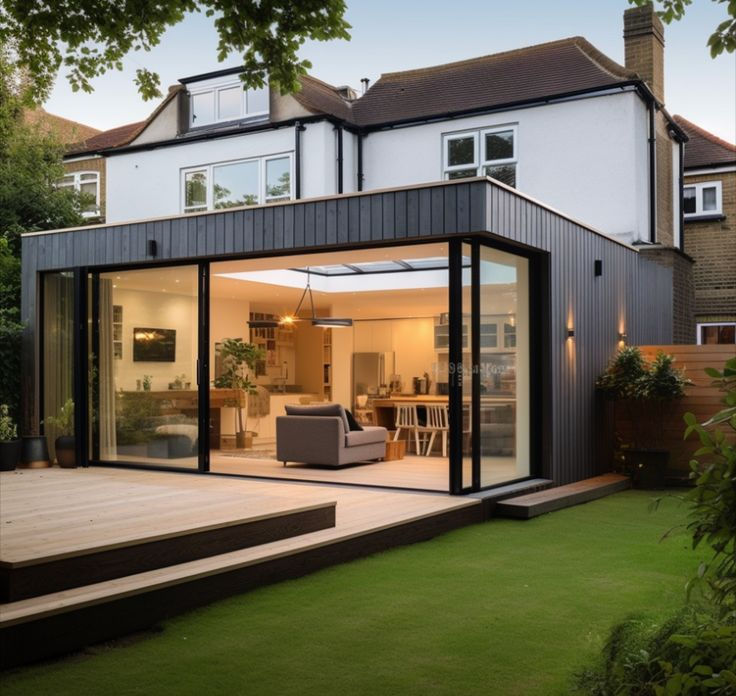 Modern house with large glass doors opens to a bright, inviting living room. Lush green lawn and wooden deck in the foreground.
