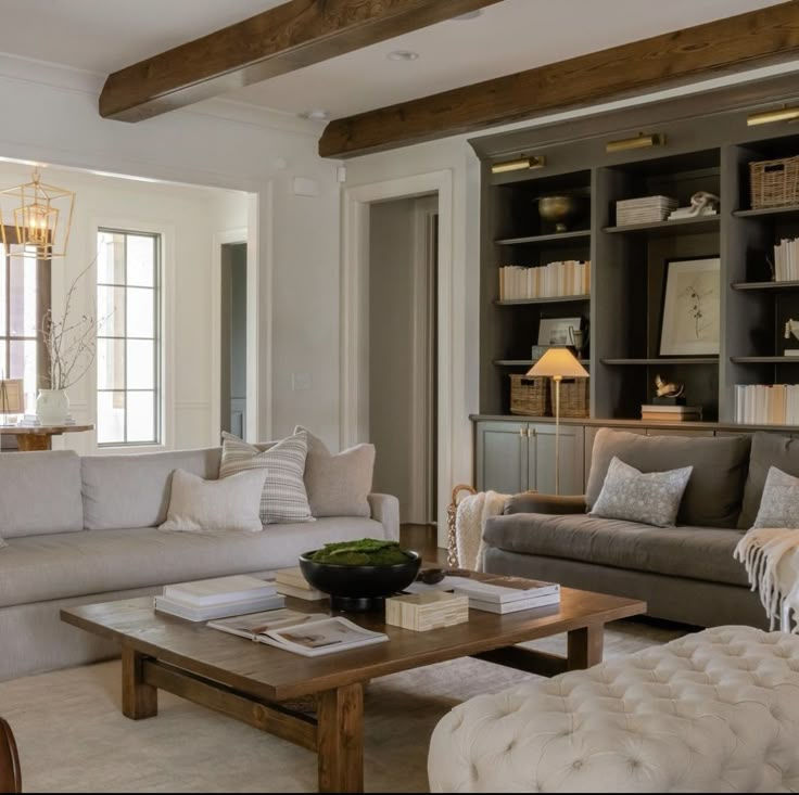 Cozy living room with beige sofas, wooden beams, and a large coffee table. Bookshelves in the background and a soft, warm ambiance.