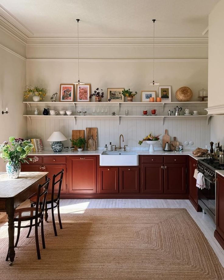 Cozy kitchen with red cabinets, a farmhouse sink, and wooden table. Shelves display plants, art, and kitchenware. Warm, inviting atmosphere.