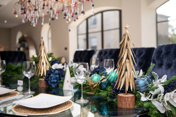 Elegant holiday table setting with gold tree centerpieces, blue ornaments, and greenery. Crystal chandelier and arched windows in the background.