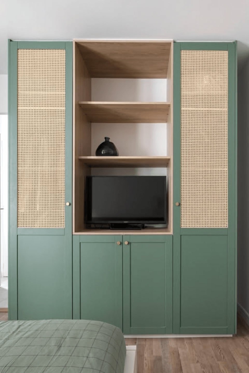 IKEA HACK - Green wardrobe with woven panels, holds TV and vase. Wooden shelves, green bed in foreground. Light, modern bedroom setting.