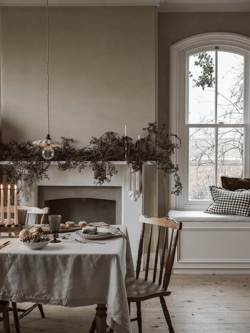 Cozy dining room with a table set for a meal, candles lit. Beige walls, window with a view, rustic decor, and a calm, inviting mood.