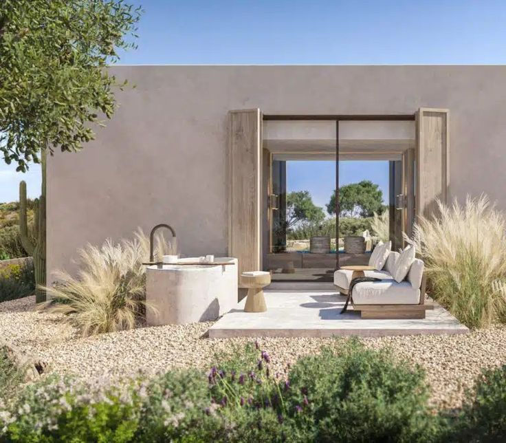 Modern patio with white cushioned chairs, round table, and cactus plants. Beige house backdrop, clear skies, and serene desert landscape.