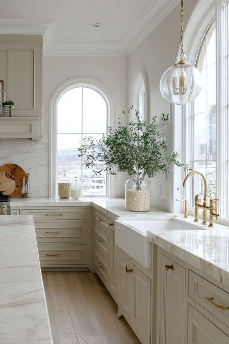 Bright kitchen with cream cabinets, marble countertops, and gold fixtures. A large window, plants, and hanging light create an airy feel.