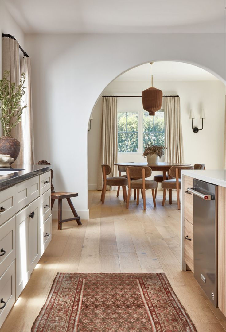 Bright kitchen with wooden floor opens to dining area. Arched entry, beige cabinets, and patterned rug. Warm, inviting atmosphere.