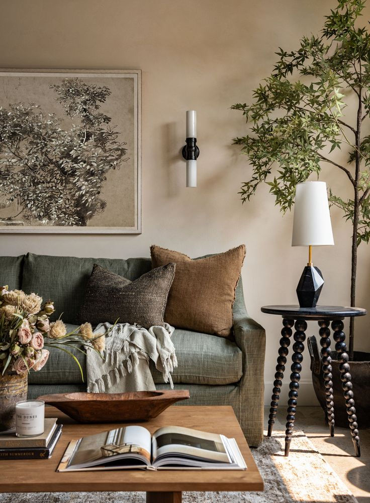 Cozy living room with green sofa, brown pillows, and a potted tree. Art on wall, books, and flowers on table. Warm, inviting ambiance.