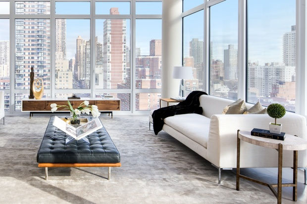 Modern living room with white sofa, black ottoman, large windows showing city skyline. Bright, elegant, with neutral tones and plants.