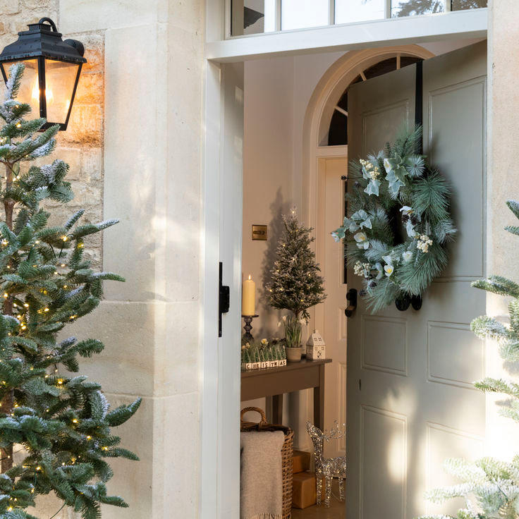 Festive doorway with a green wreath, lit outdoor trees, and a decorated interior with candles and a mini tree, creating a cozy atmosphere.