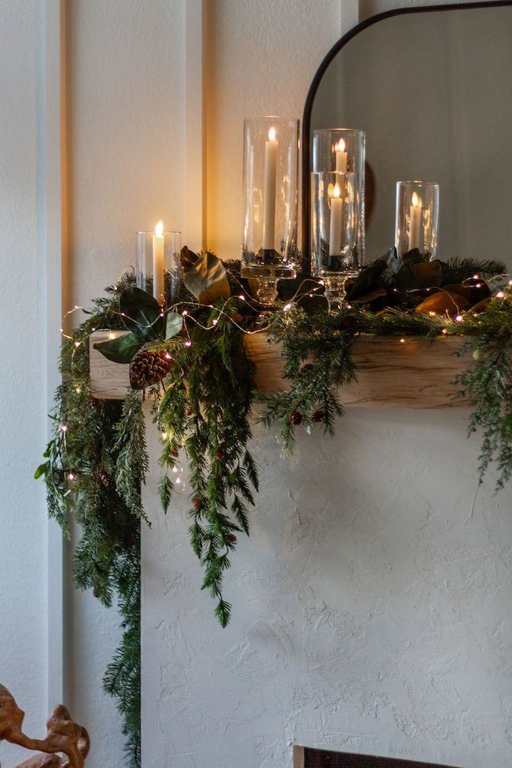 Candles in glass holders on a mantel with pine garland and string lights. Soft illumination creates a warm, festive atmosphere.