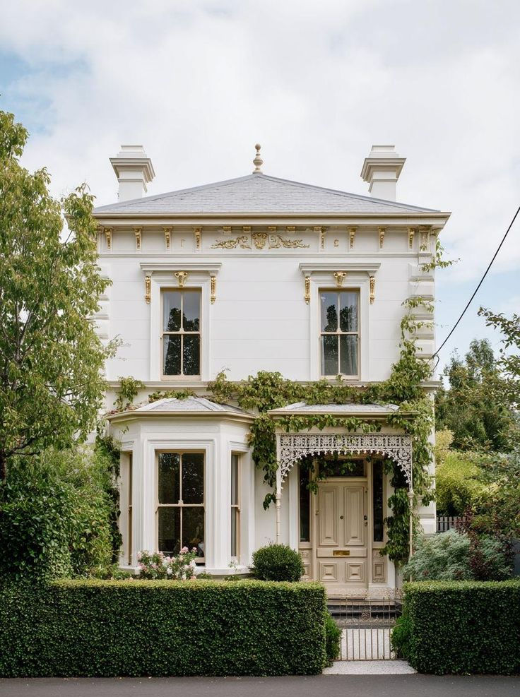 Classic two-story white house with green vines, ornate gold details, and manicured hedges, under a cloudy sky. Text: "everhomestyle.com."