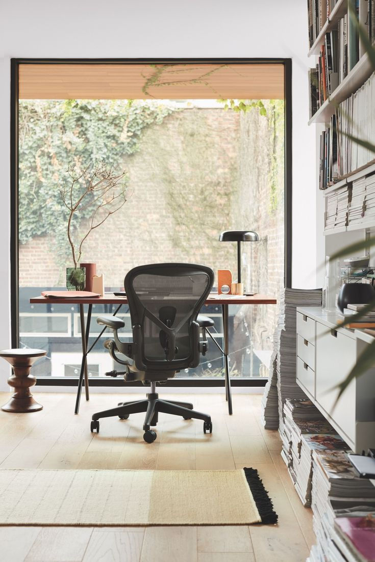 Office with a black chair facing a desk, situated by a large window showing an ivy-covered wall. Magazines stacked on right. Calm ambiance.