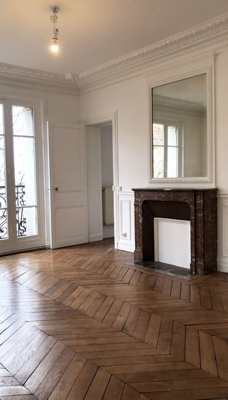 Empty room with herringbone wood floor, white walls, ornate ceiling, large mirror over marble fireplace, and French doors opening to balcony.
