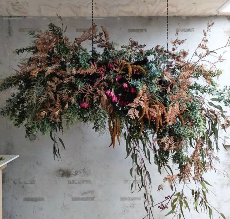 Hanging floral arrangement with green leaves, brown ferns, and pink flowers against a textured gray wall. Rustic and natural ambiance.