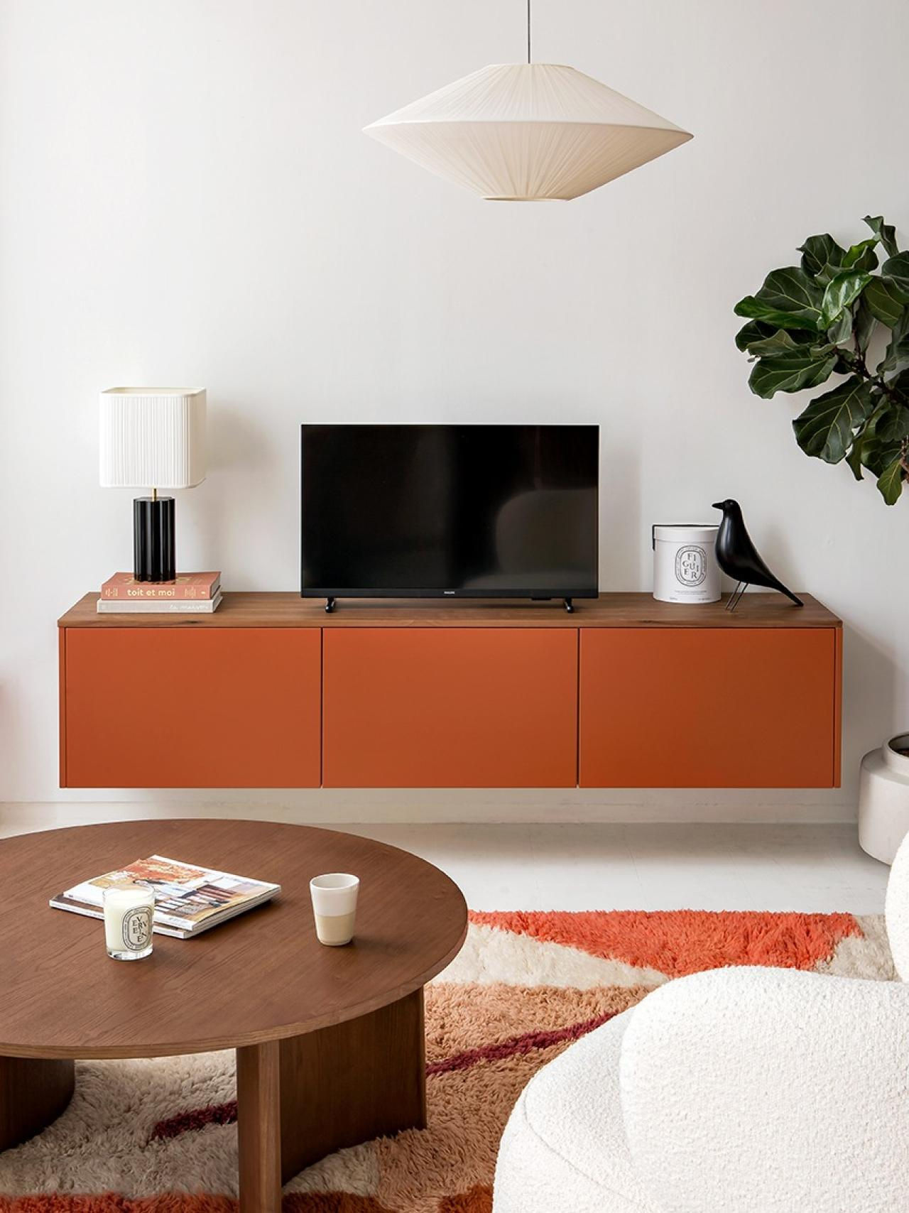 IKEA HACK Minimalist living room with orange cabinet, TV, black bird figurine, and lamp. Round table with magazines, candle, and cup. Cozy vibe.