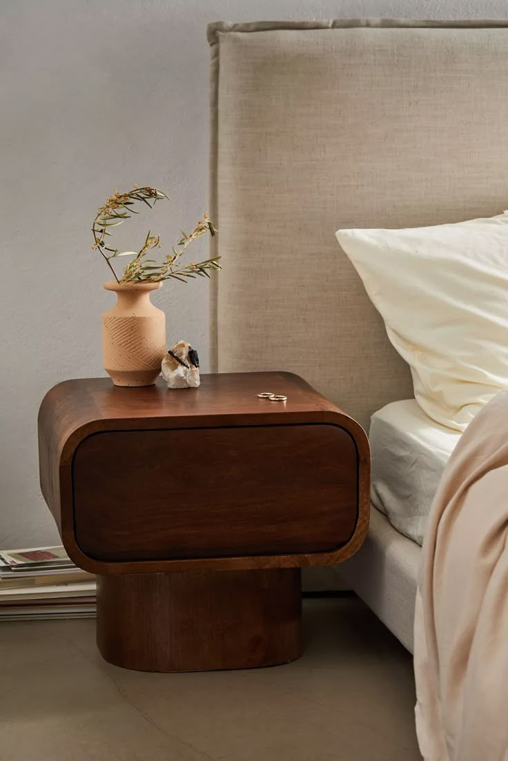 Wooden bedside table with vase and dried branches, crystals, and rings. Beige bed, soft lighting, creating a serene bedroom mood.