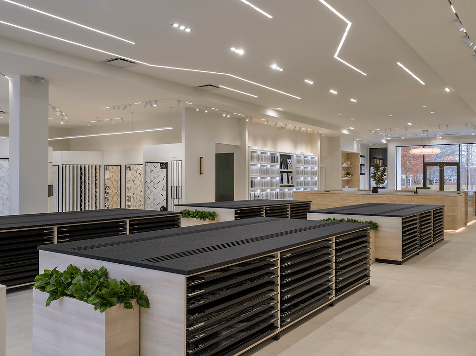Spacious tile showroom with sleek tables and green plants. Backlit display shelves, modern lighting, and large windows create a bright setting.