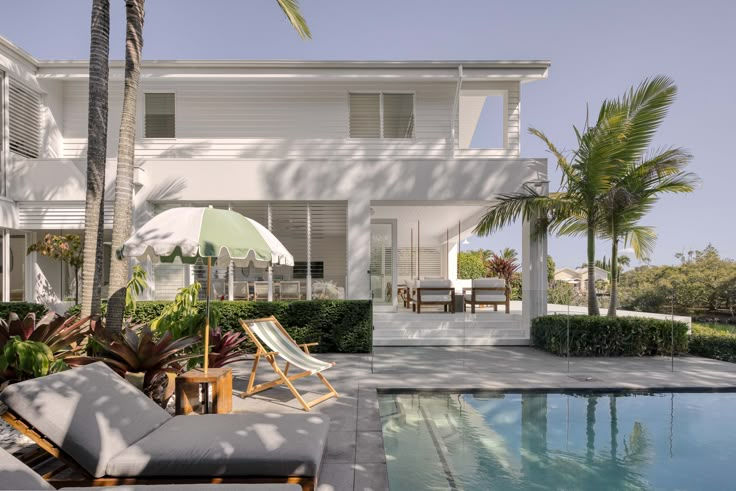 Modern white house with pool, palm trees, and lounge chairs under a green and white umbrella. Sunny, tranquil outdoor setting.