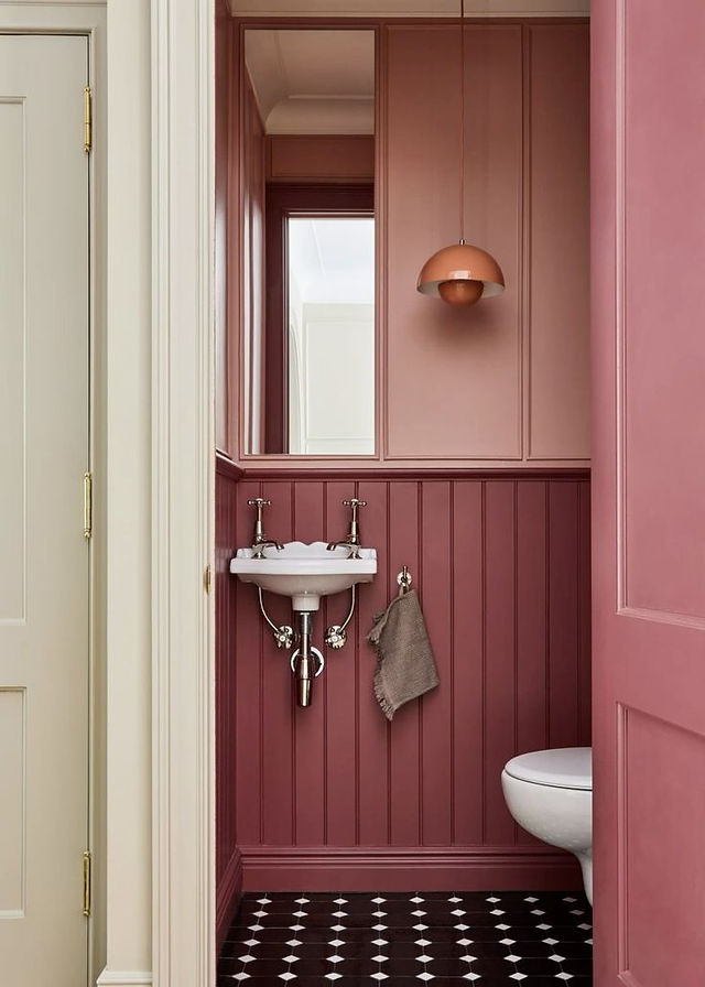 Elegant pink bathroom with a round mirror, white pedestal sink, brass fixtures, and checkered floor. A towel hangs on the sink.