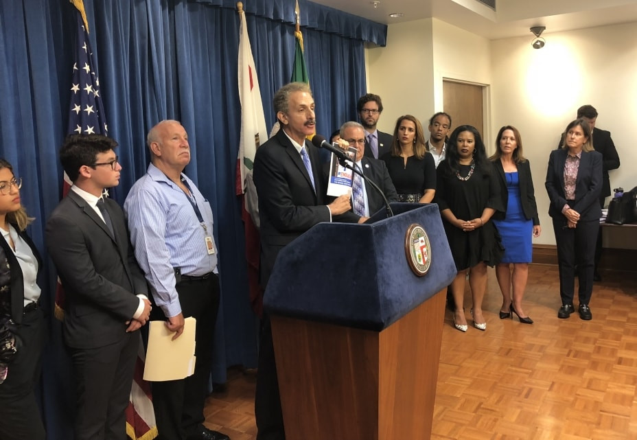 City Attorney Mike Feuer at podium holding a copy of the Report of the Blue Ribbon Panel on School Safety, flanked by members, City Attorney staff members and invited guests.