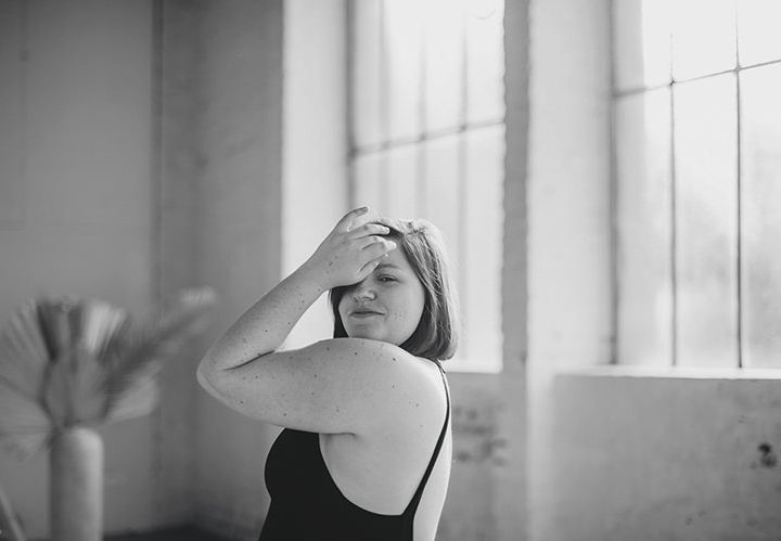 In dieser Boudoir-Fotografie von Mira Mikosch sehen wir eine intime Schwarz-Weiß-Aufnahme einer Frau, die in einem minimalistischen Raum steht, in dem das Licht durch große Fenster strömt. Das Model ist mit einem schlichten schwarzen Top bekleidet, das die Schultern freilegt, und sie dreht sich leicht zur Kamera, während sie eine Hand in ihrem Haar hat. Ihr Gesichtsausdruck strahlt Selbstbewusstsein und eine gewisse Verspieltheit aus, was die natürliche Schönheit und Sinnlichkeit des Moments unterstreicht. Die weiche Beleuchtung und der Hintergrund aus Fenstern und Pflanzen tragen zur ruhigen und eleganten Atmosphäre bei, die typisch für Mira Mikoschs Boudoir-Fotografie ist.