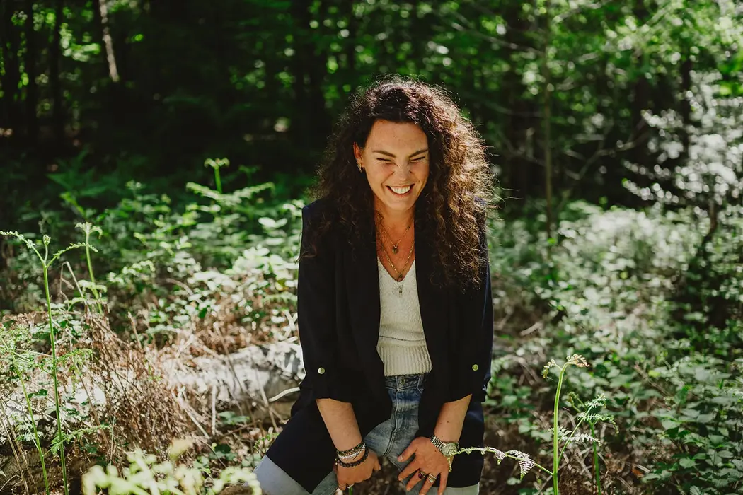 Frau lacht herzlich im Wald und beugt sich nach vorne zwischen grünen Pflanzen.