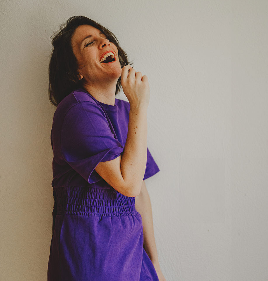 Porträt von Mira Mikosch, Personal Branding und Boudoir-Fotografin aus Mönchengladbach, NRW. Sie trägt ein lila T-Shirt und lacht herzlich, während sie sich entspannt an einer Wand anlehnt. Ihre offene und authentische Ausstrahlung wird durch das spontane Lächeln betont.