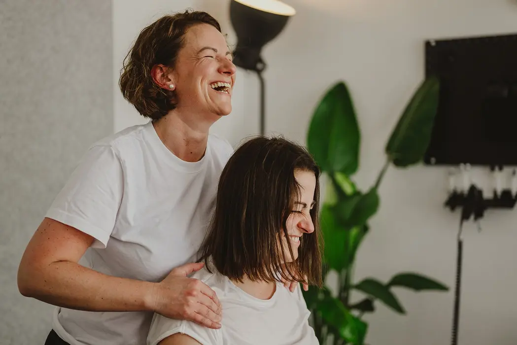 Zwei Frauen lachen während einer entspannten chiropraktischen Behandlung in Praxisraum mit Pflanzen.