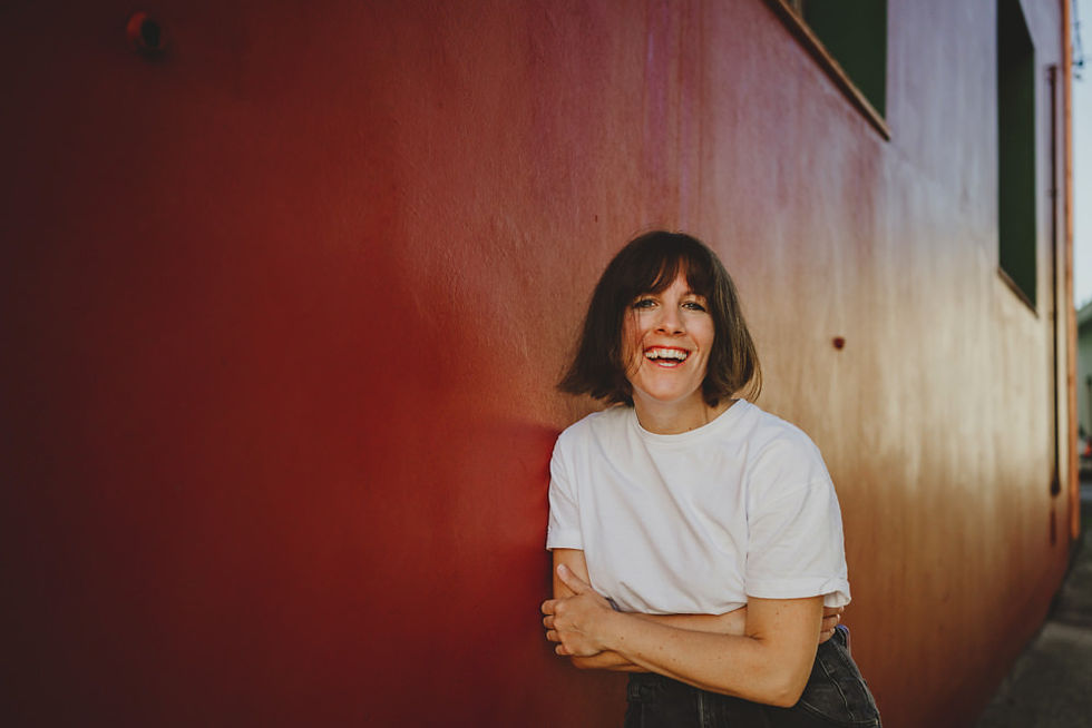 Eine Frau steht an einer roten Wand und schaut lachend in die Kamera.