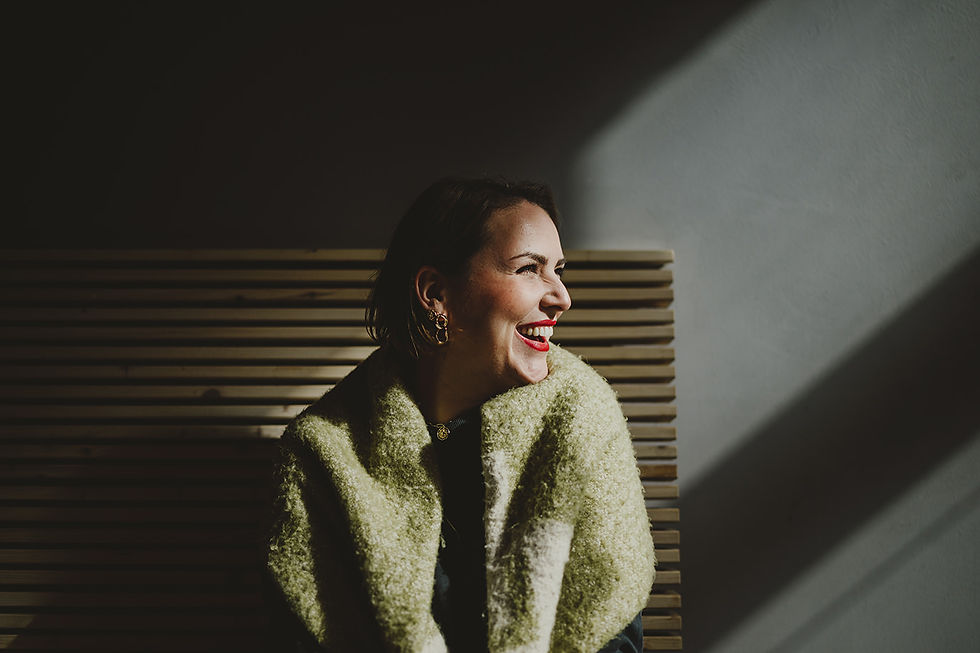 Eine Frau mit kurzen Haaren und kräftigem roten Lippenstift sitzt vor einer hölzernen, gestreiften Wand und lacht herzlich zur Seite. Sie trägt goldene Ohrringe und einen warmen, grünen Schal. Dieses Personal Branding Foto von Mira Mikosch kombiniert Studiofotografie mit kreativem Lichtspiel und vermittelt Eleganz und Lebensfreude.
