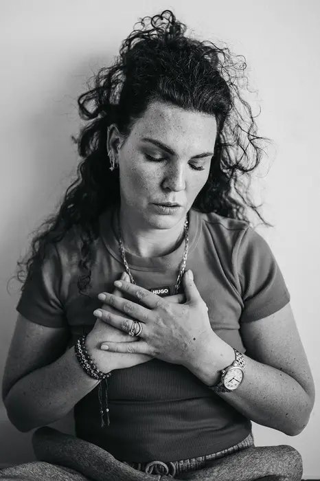 Frau sitzt meditativ mit geschlossenen Augen und Händen auf dem Herzen vor heller Wand.