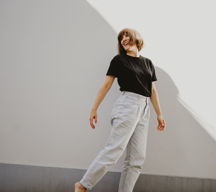 Bild von Mira Mikosch, Personal Branding und Boudoir-Fotografin aus Mönchengladbach, NRW. Sie trägt ein schwarzes T-Shirt und weiße Jeans, während sie sich in einem lockeren Moment an einer Wand entlang dreht und dabei lacht. Das Bild strahlt eine fröhliche, lebendige Energie aus und zeigt ihre entspannte, authentische Persönlichkeit.
