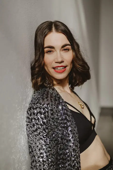 Porträt einer lächelnden Frau mit glitzerndem Blazer und schwarzem Oberteil an einer Wand.