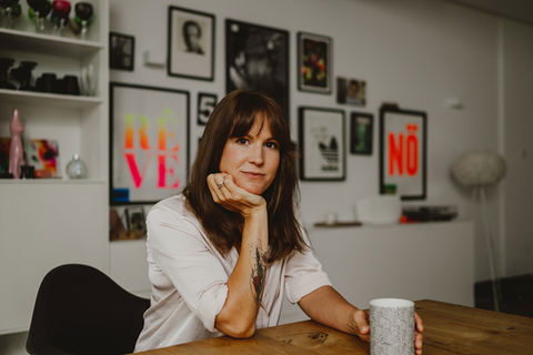 Kreative Unternehmerin sitzt entspannt am Schreibtisch in modernem Studio und lächelt in die Kamera – authentische Businessfotografie mit persönlicher Ausstrahlung.