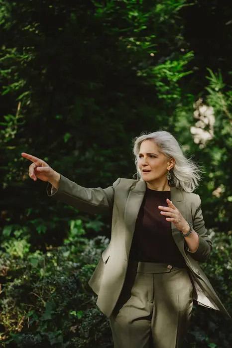 Frau im grünen Hosenanzug zeigt gestikulierend in die Ferne inmitten eines Parks.