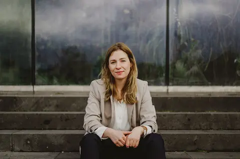 Frau sitzt entspannt vor moderner Glasfassade im Freien – natürliche Businessfotografie mit urbanem Look und authentischer Ausstrahlung.