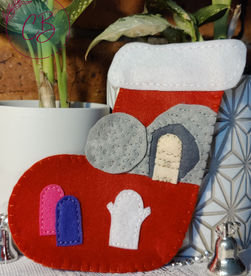 Red felt Christmas stocking with stitched scene involving a cave and colourful figures, set against a brick and plant background. Silver bells sit nearby.