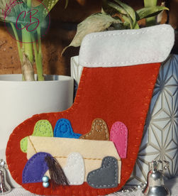 Red felt Christmas stocking with colorful felt decorations on a table, plants in background. Silver bells and white pots add festive touch.