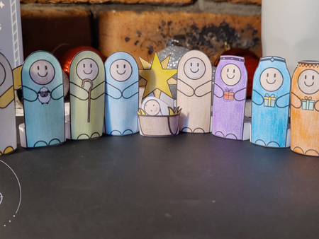 Colourful Nativity finger puppets crafted from paper standing in a row on a dark foreground. 