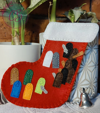 Red felt stocking with colourful felt shapes attached, set against potted plants on a textured white surface. Small silver bell nearby.